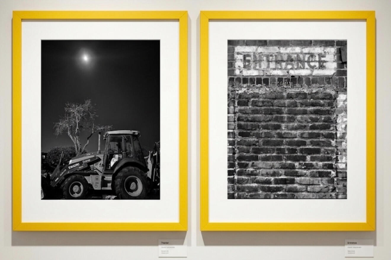 tractor & entrance at TAG Gallery Two black-and-white framed photographs hanging side-by-side on a gallery wall. The left photograph depicts a tractor in a field at night under a bright moon and a leafless tree. The right photograph shows a weathered brick wall with a rectangular sign abov